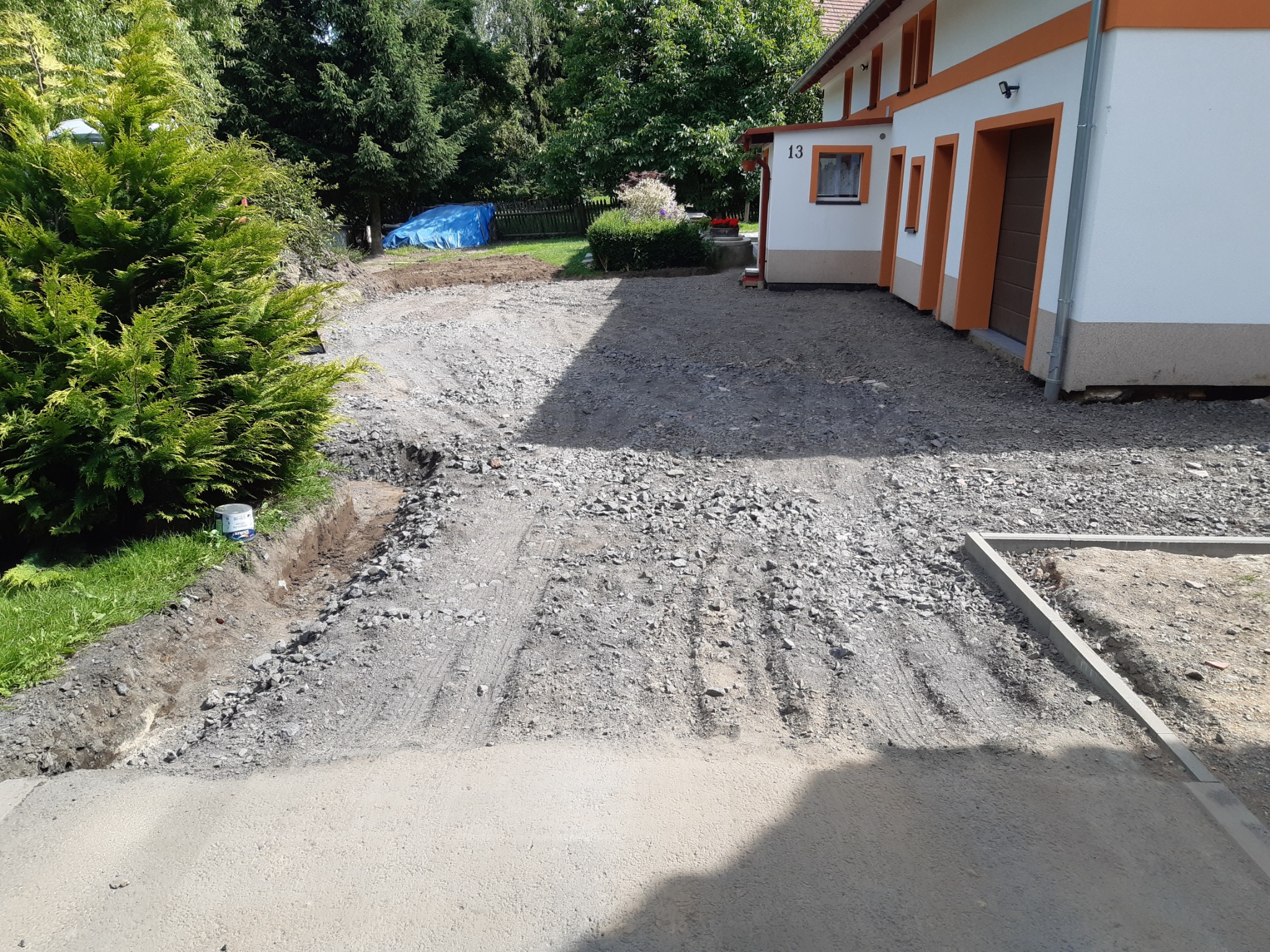 Przygotowanie podłoża pod kostkę brukową na posesji, widoczne obrzeża, warstwa tłucznia i piasku, w tle budynek z numerem 13 i zieleń.