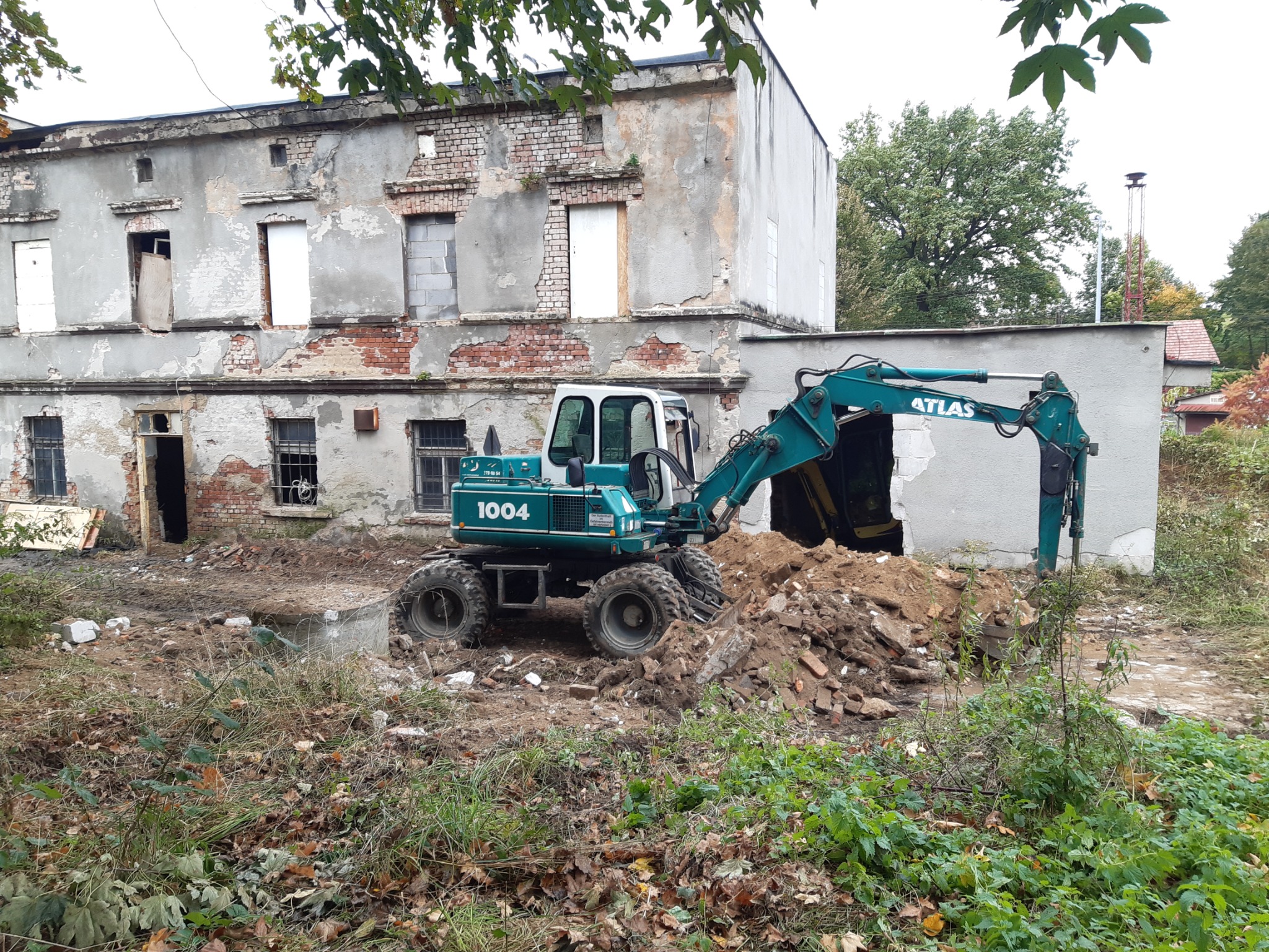 Zielona koparka Atlas 1004 na tle częściowo rozebranego, dwukondygnacyjnego budynku z cegły i betonu, z widocznymi otworami okiennymi i drzwiowymi, otoczona gruzem i roślinnością.