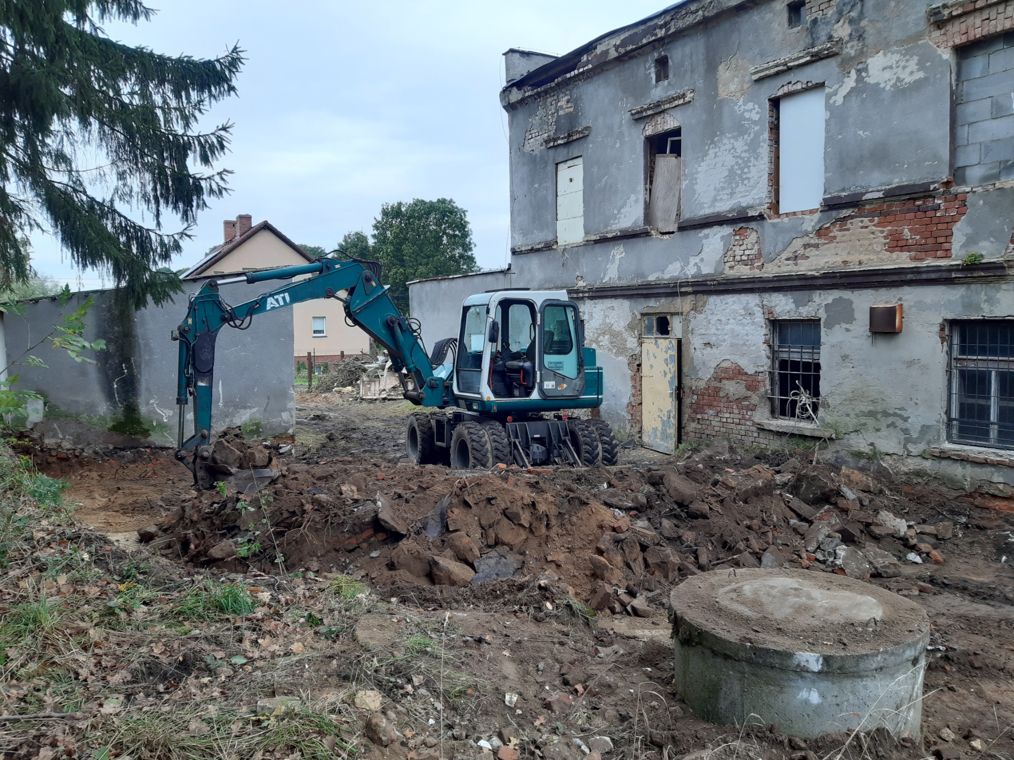 Niebieska koparka kołowa ATI podczas prac ziemnych na placu budowy z gruzem i fragmentami cegieł, na tle zniszczonego budynku z wybitymi oknami i zamurowanymi drzwiami.