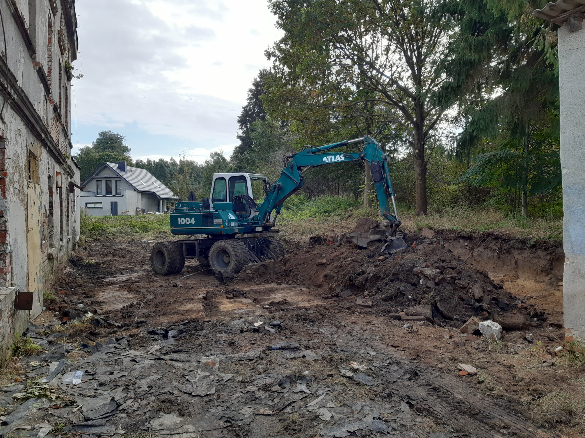 Niebieska koparka Atlas 1004 w trakcie prac ziemnych na terenie budowy, z widocznymi fragmentami gruzu i starego budynku w tle.