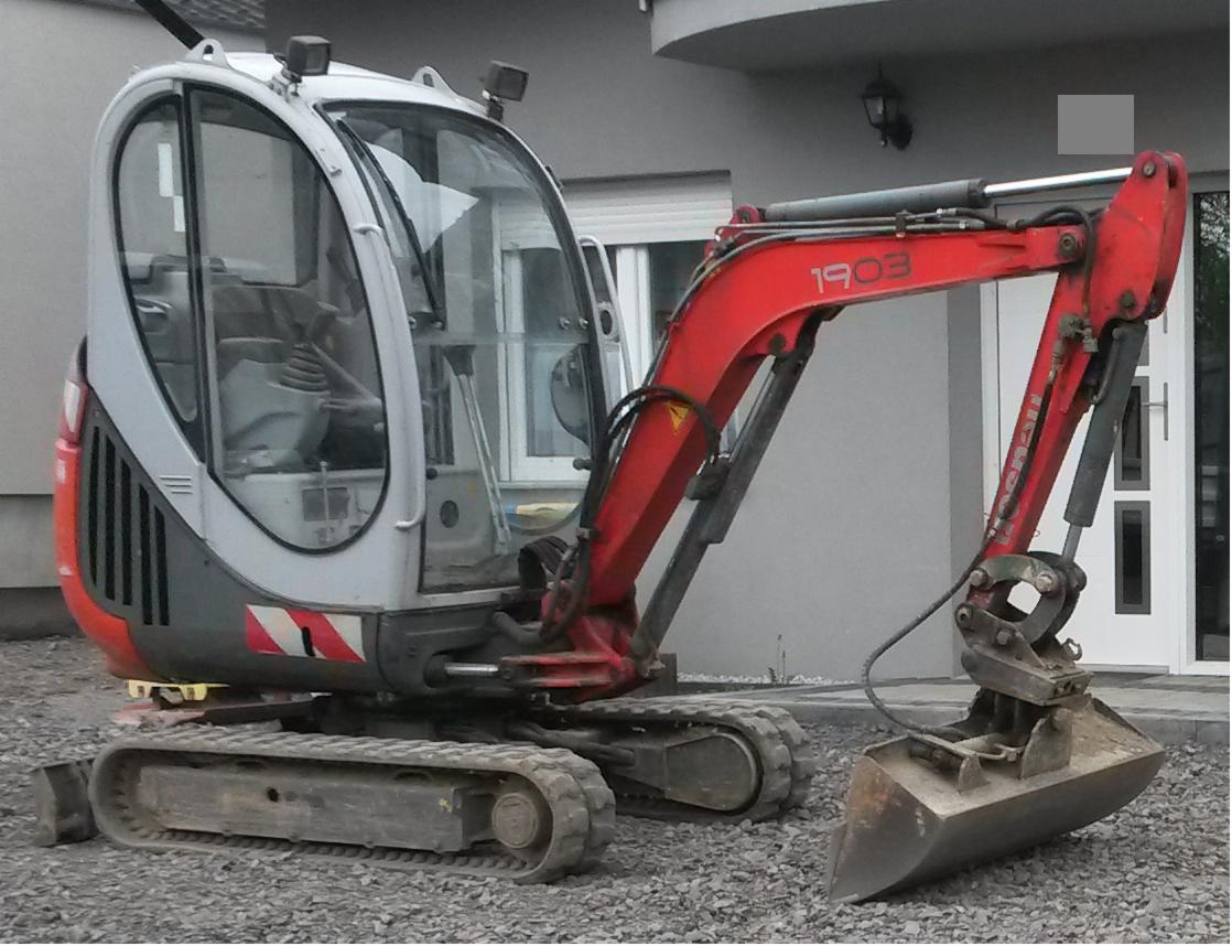 Kompaktowa, czerwono-szara mini koparka na gąsienicach, zaparkowana na żwirowym podłożu przed budynkiem. Widoczny wysięgnik z łyżką i kabina operatora.