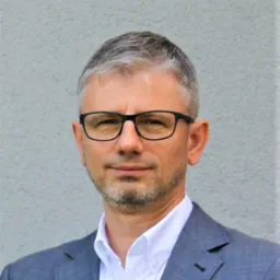 Portret mężczyzny w okularach i szarej marynarce, z siwiejącymi włosami, na tle szarej ściany.