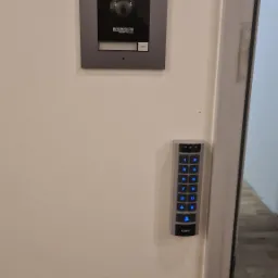 Zamontowany na ścianie panel domofonu z kamerą i klawiaturą numeryczną z podświetlanymi na niebiesko cyframi.