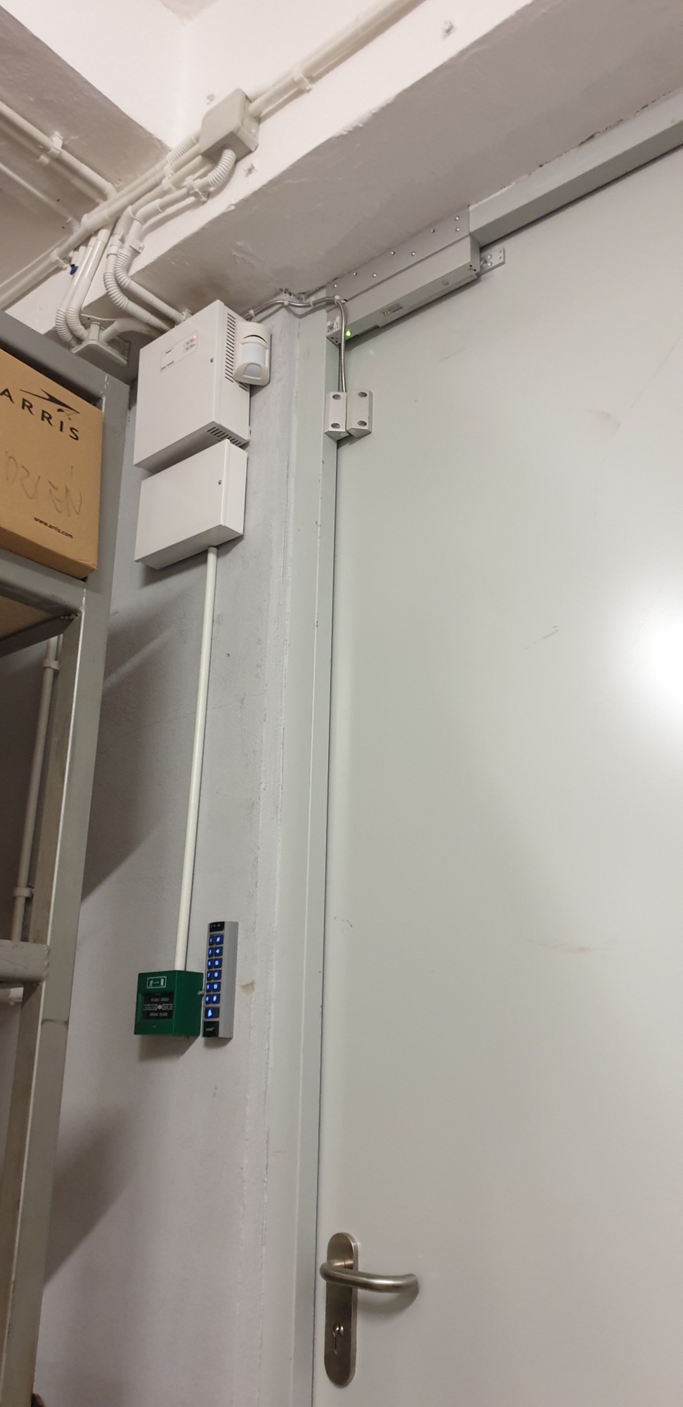 Białe drzwi z zamontowanym systemem kontroli dostępu: panel z klawiaturą numeryczną, czytnik kart i elektrozaczep na ścianie, instalacja elektryczna widoczna na suficie.