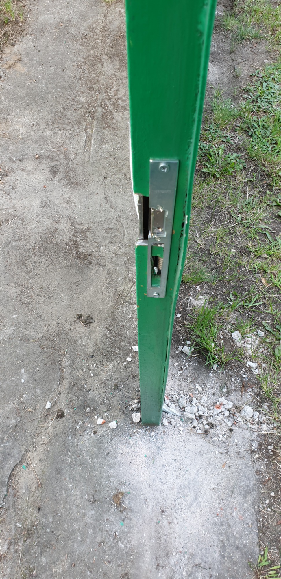 Zielony słupek z uszkodzonym zamkiem, fragment betonowego podłoża i trawy.