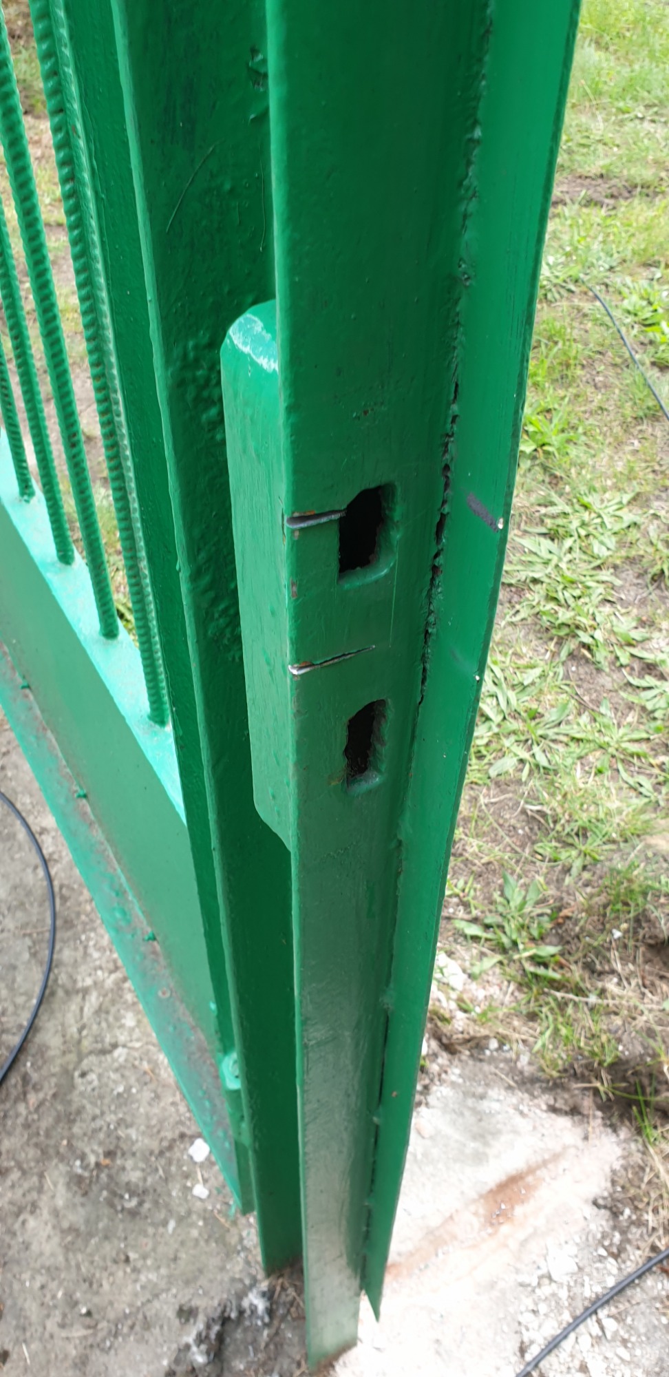 Fragment zielonej metalowej bramy z dwoma kwadratowymi otworami, widoczne ślady spawania i niedokładnego malowania, tło z trawy i betonu.