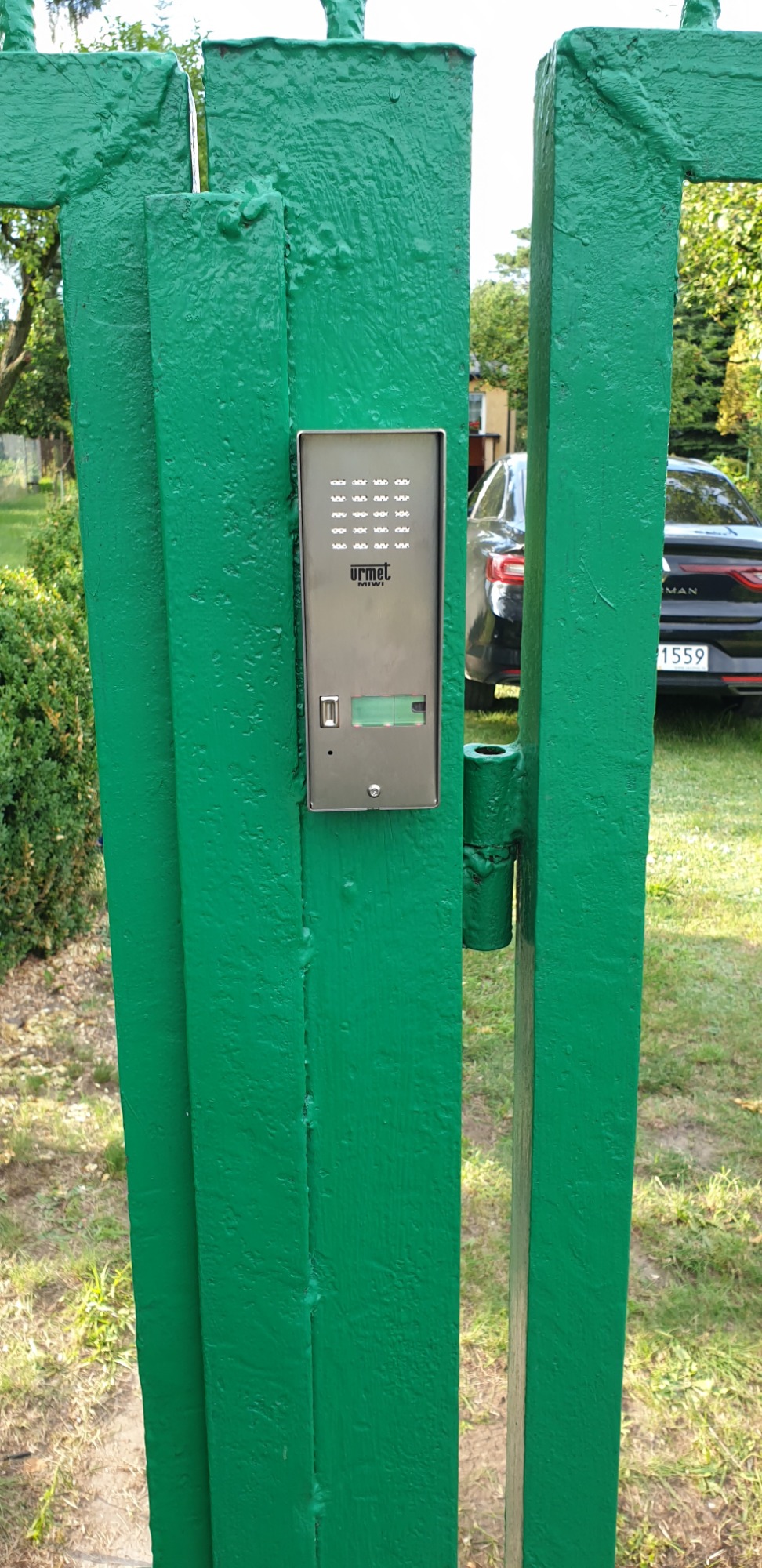 Zielona, pomalowana brama wejściowa z zamontowanym panelem domofonu Urmet Miwi, widoczny fragment zaparkowanego samochodu w tle.
