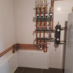 Kompleksowa instalacja grzewcza z miedzianymi rurami, zaworami i pompami obiegowymi zamontowana na ścianie wyłożonej białymi płytkami, z widocznym zbiornikiem wyrównawczym i szafką rewizyjną...