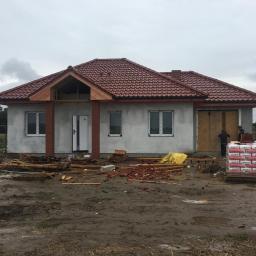 Intimo - Surowy stan domu murowanego z czerwoną dachówką, widoczne ceglane filary wejściowe i sterta zapakowanych materiałów budowlanych na błotnistym terenie.