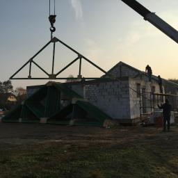 Intimo - Montaż konstrukcji dachowej z użyciem dźwigu na budowie domu z bloczków, widoczni pracownicy na rusztowaniu i gotowe elementy więźby dachowej.