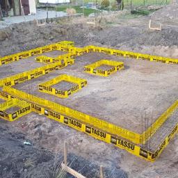 Intimo - Przygotowanie fundamentów pod budynek jednorodzinny, widoczne szalunki systemowe z żółtymi obudowami i zbrojenie, teren budowy z wykopem i usypaną ziemią w tle.