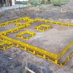 Przygotowanie fundamentów pod budynek jednorodzinny, widoczne szalunki systemowe z żółtymi obudowami i zbrojenie, teren budowy z wykopem i usypaną ziemią w tle.