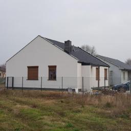 Intimo - Nowoczesny, parterowy dom z białymi ścianami i ciemną dachówką, z brązowymi roletami w oknach, otoczony siatką ogrodzeniową i niezagospodarowanym terenem.