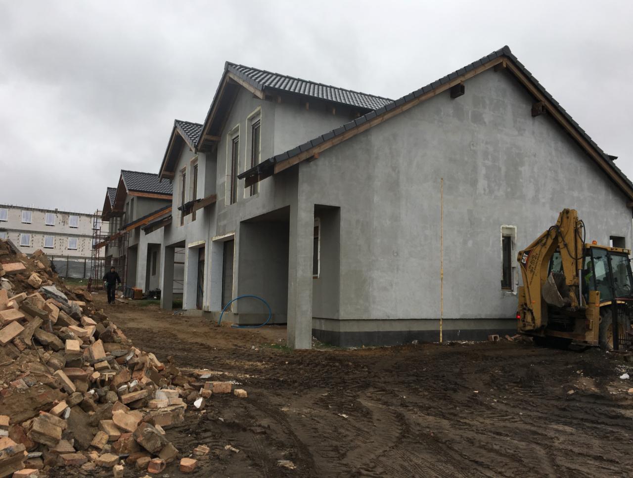 Szeregówka domów w stanie surowym zamkniętym z szarym tynkiem zewnętrznym, widoczny stos cegieł i koparka CAT na placu budowy w pochmurny dzień.