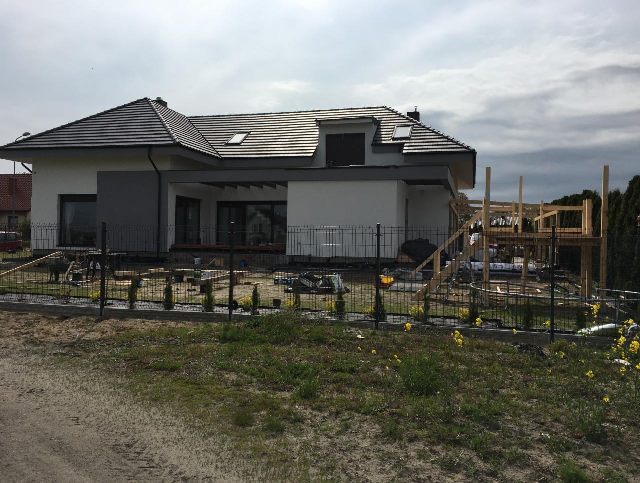 Wznoszony dom jednorodzinny z ciemną dachówką i elementami szarości, widoczne drewniane elementy konstrukcji tarasu i przygotowany teren pod ogród.