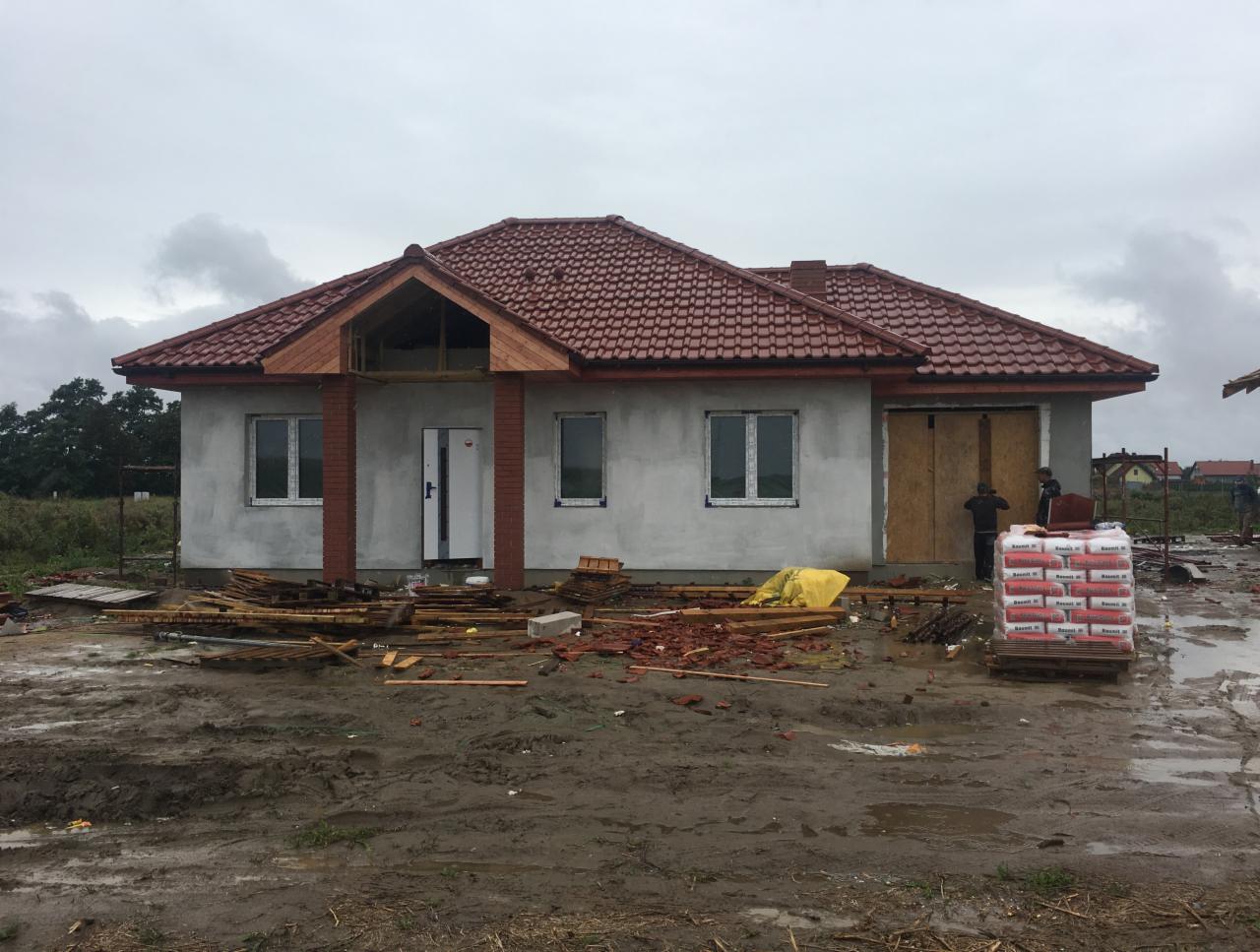 Surowy stan domu murowanego z czerwoną dachówką, widoczne ceglane filary wejściowe i sterta zapakowanych materiałów budowlanych na błotnistym terenie.