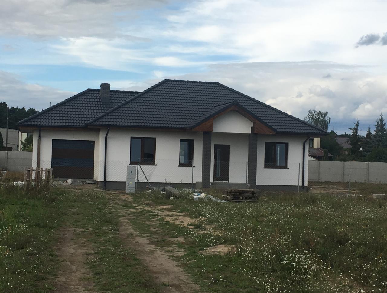 Nowo wybudowany parterowy dom jednorodzinny z ciemną dachówką, białymi ścianami i garażem, otoczony niezagospodarowanym terenem z widocznymi śladami budowy i paletami.