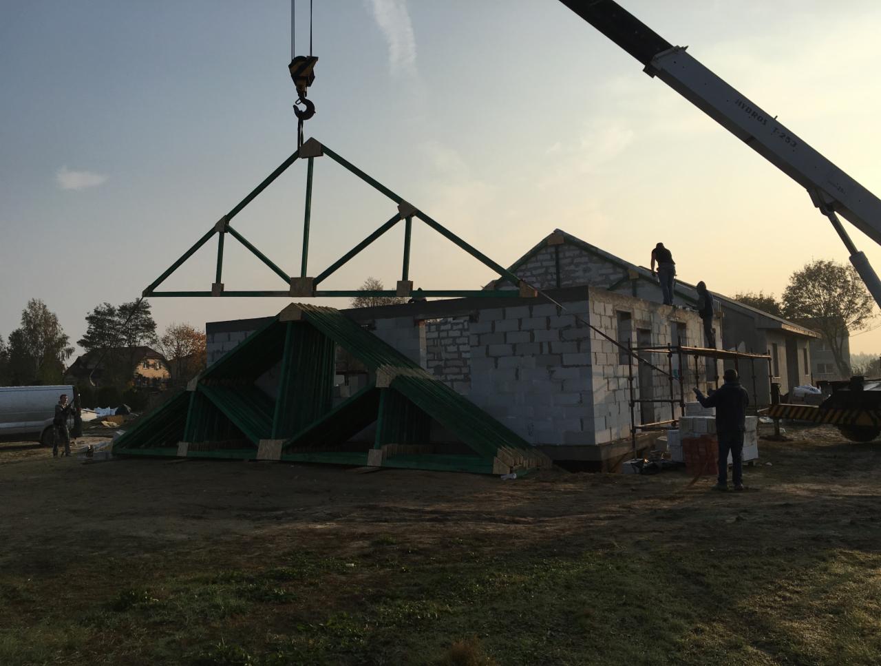 Montaż konstrukcji dachowej z użyciem dźwigu na budowie domu z bloczków, widoczni pracownicy na rusztowaniu i gotowe elementy więźby dachowej.