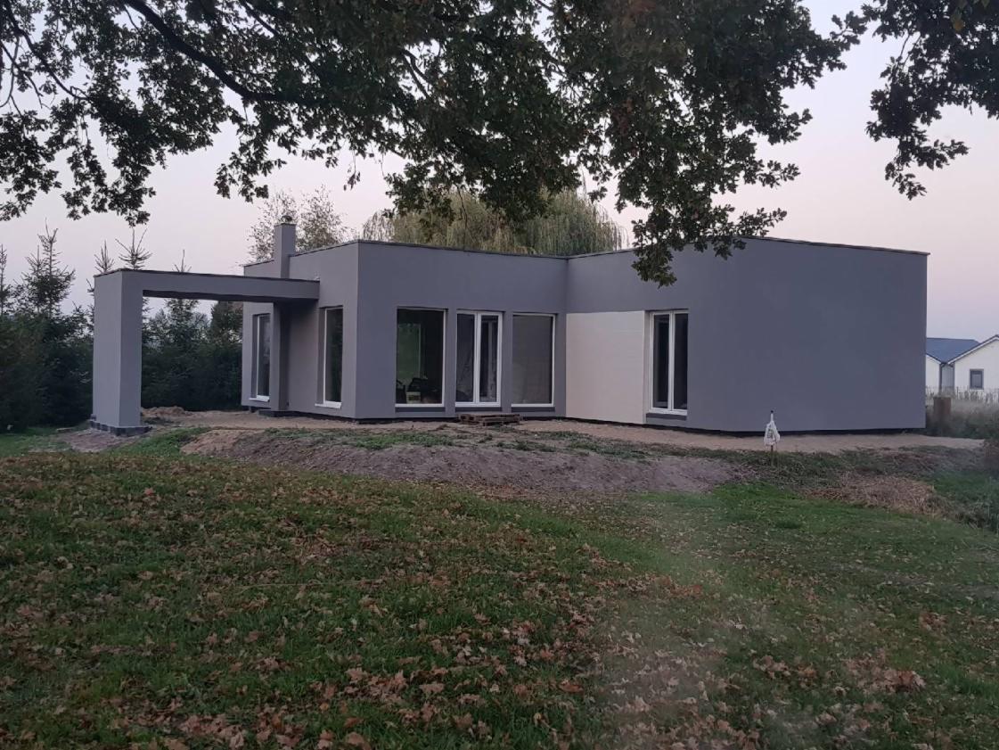 Nowoczesny dom jednorodzinny z płaskim dachem, wykończony w odcieniach szarości i bieli, z dużymi oknami i zadaszonym wejściem, otoczony trawnikiem i drzewami.