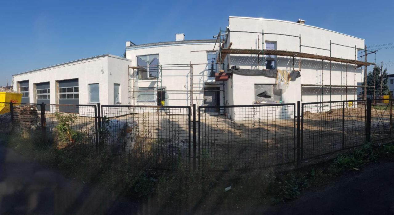 Dwukondygnacyjny budynek z płaskim dachem w trakcie remontu elewacji, widoczne rusztowania, okna zabezpieczone folią, teren ogrodzony metalową siatką.