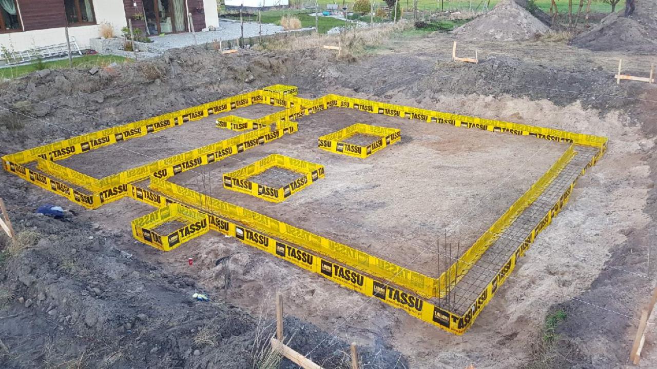 Przygotowanie fundamentów pod budynek jednorodzinny, widoczne szalunki systemowe z żółtymi obudowami i zbrojenie, teren budowy z wykopem i usypaną ziemią w tle.