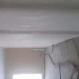 Surowy pokój w trakcie wykańczania z widoczną świeżo nałożoną szarą gładzią na ścianach i suficie, wystające kable elektryczne i widoczne niedoskonałości powierzchni.