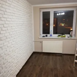 Pokój po remoncie: ściana wyłożona dekoracyjną białą cegłą, ciemna drewniana podłoga, biały grzejnik pod oknem z widokiem na nocne miasto.