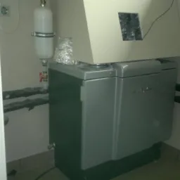 Szaro-zielona centrala wentylacyjna z podłączonymi rurami w pomieszczeniu technicznym, widoczny biały zbiornik wyrównawczy i fragment instalacji elektrycznej.