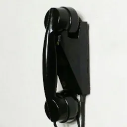 Czarny, zabytkowy telefon ścienny z kablem, zamontowany na białej ścianie, widok z boku.