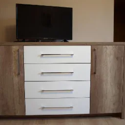 Komoda z czterema białymi szufladami i dwojgiem drzwi z okleiną imitującą jasne drewno, na której stoi telewizor.