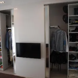 Biała szafa wnękowa z przesuwnymi drzwiami lustrzanymi, wbudowany telewizor na ścianie, półki z ubraniami i akcesoriami.