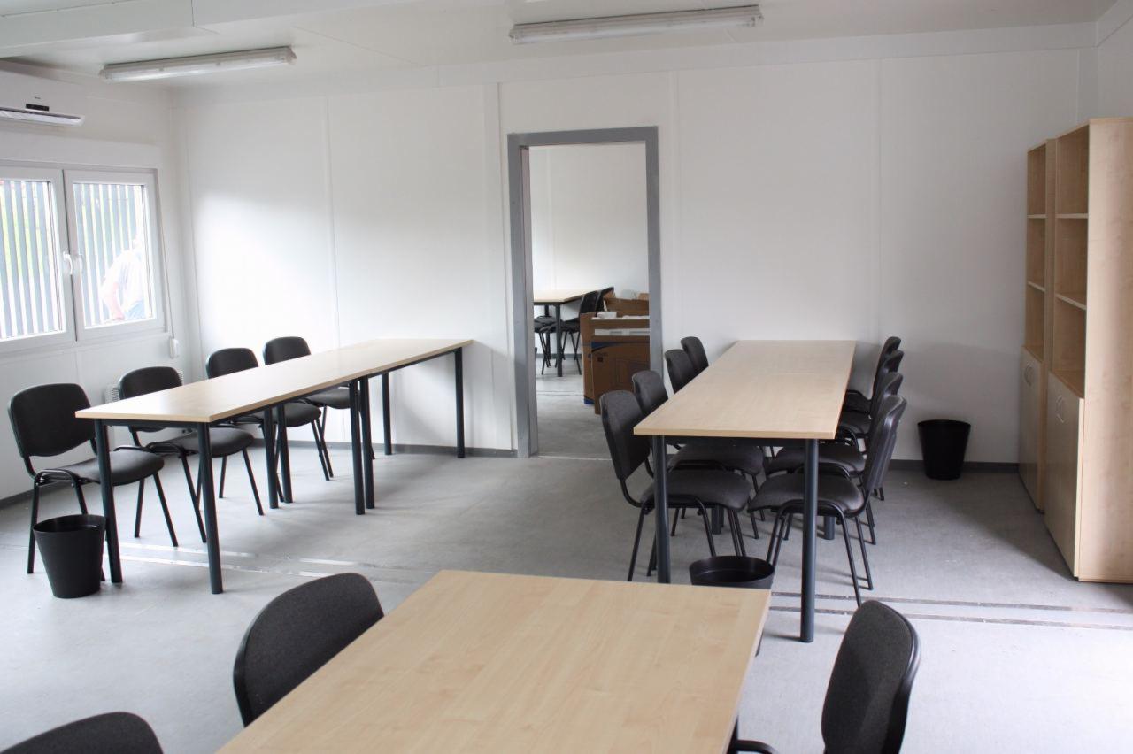 Jasne biuro z trzema długimi, drewnianymi stołami z czarnymi nogami i krzesłami, widok na kolejne pomieszczenie przez otwarte drzwi.