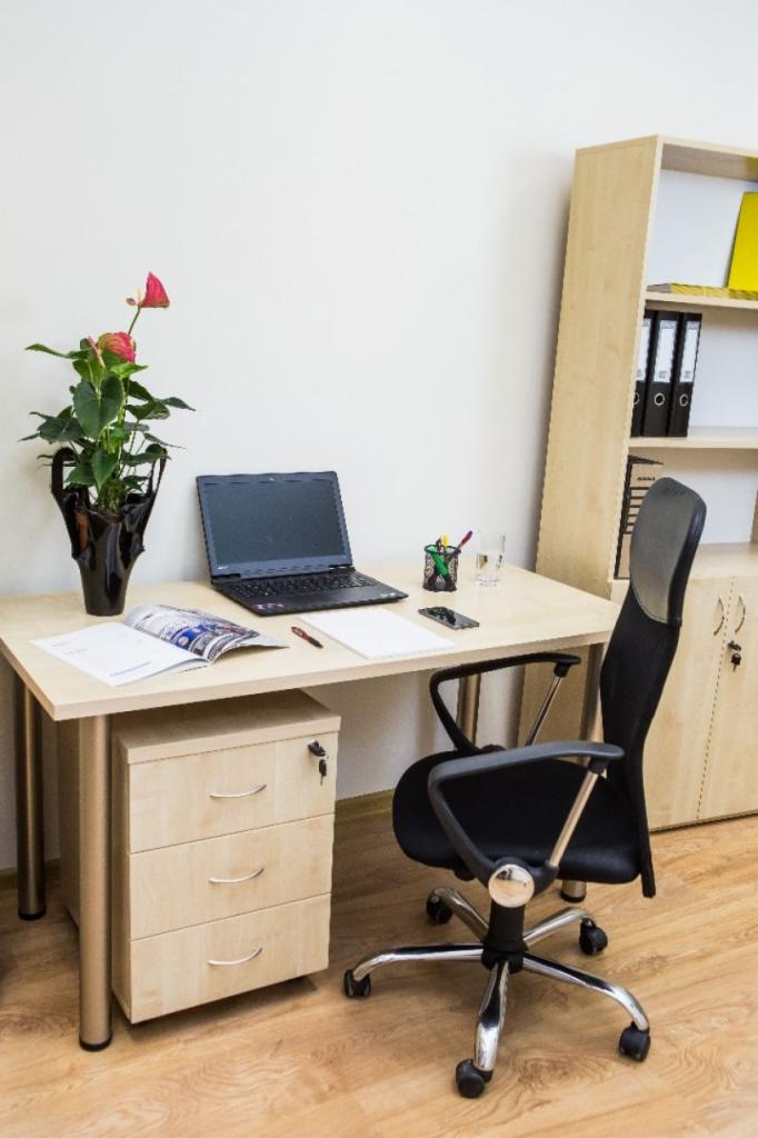 Biurko z blatem w kolorze jasnego drewna, z trzema szufladami po lewej stronie, czarnym krzesłem obrotowym, laptopem, kwiatem anturium w czarnym wazonie i regałem w tle.