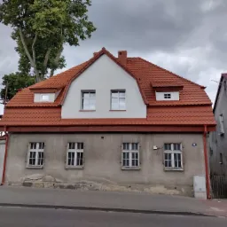 Dwupiętrowy dom z nowym, czerwonym dachem z dachówki karpiówki, szarymi ścianami i białymi oknami, widok z ulicy w pochmurny dzień.