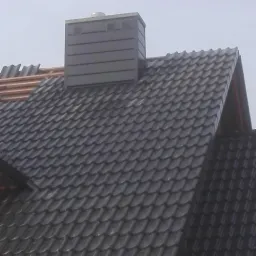 Fragment dachu z czarnej dachówki karpiówki i kominem z blachy w kolorze antracytowym, widoczne elementy drewnianej konstrukcji dachu w trakcie prac.