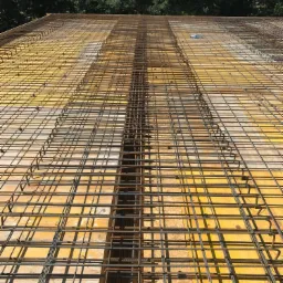 Zbrojenie stropu domu jednorodzinnego ułożone na żółtym szalunku, widok z góry na tle zieleni drzew.