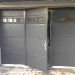 Częściowo otwarta, segmentowa brama garażowa w odcieniach szarości z poziomymi przetłoczeniami i przeszkleniami, widoczna z zewnątrz na tle kostki brukowej.