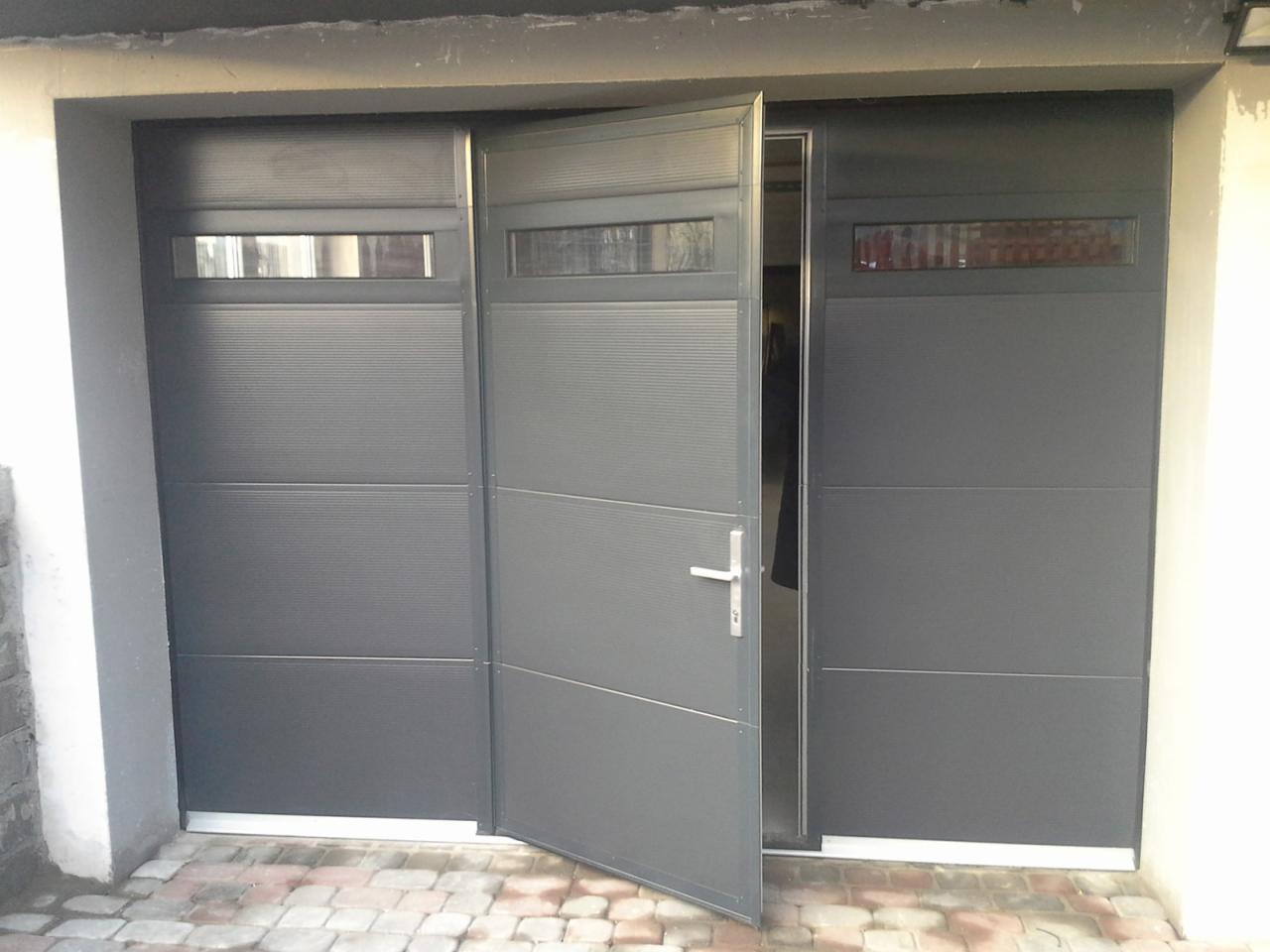 Częściowo otwarta, segmentowa brama garażowa w odcieniach szarości z poziomymi przetłoczeniami i przeszkleniami, widoczna z zewnątrz na tle kostki brukowej.