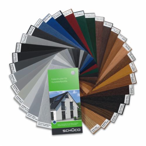 Paleta kolorów oklein do profili okiennych i drzwiowych SCHÜCO, prezentująca szeroki wybór odcieni i tekstur, od szarości i bieli po różne rodzaje drewna, z broszurą reklamową na pierwszym planie.
