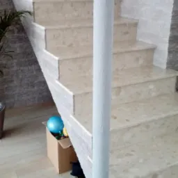 Marmurowe schody wewnętrzne z szarą kolumną wspierającą, widoczna ściana wykończona kamieniem dekoracyjnym i podłoga z paneli, w tle karton z zabawkami.