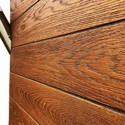 Fragment elewacji domu z brązowych, poziomych desek imitujących drewno, oświetlony lampą zewnętrzną, widoczny narożnik budynku.