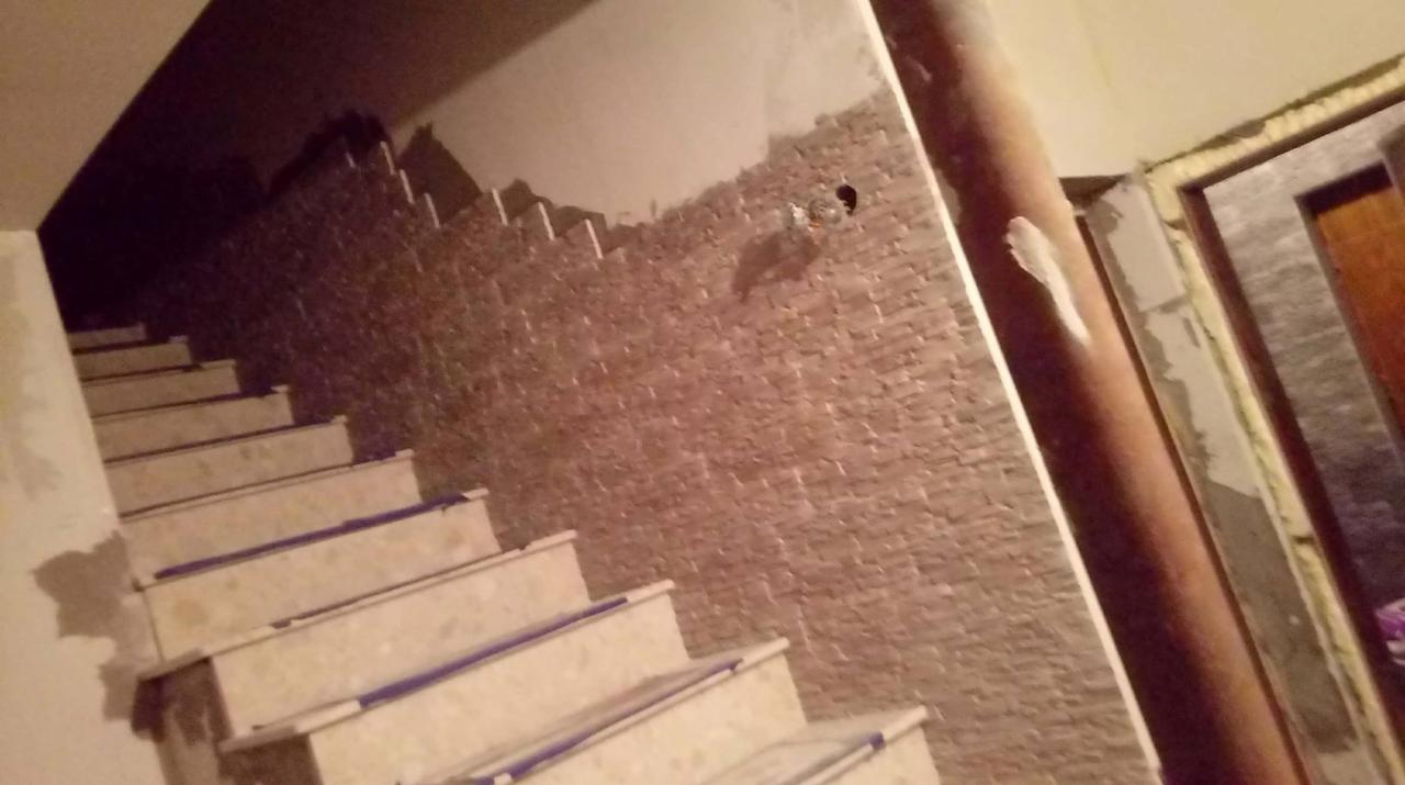 Wnętrze w trakcie remontu: schody wyłożone płytkami, ściana z dekoracyjną okładziną kamienną, widoczne niedokończone wykończenia przy futrynie drzwi.