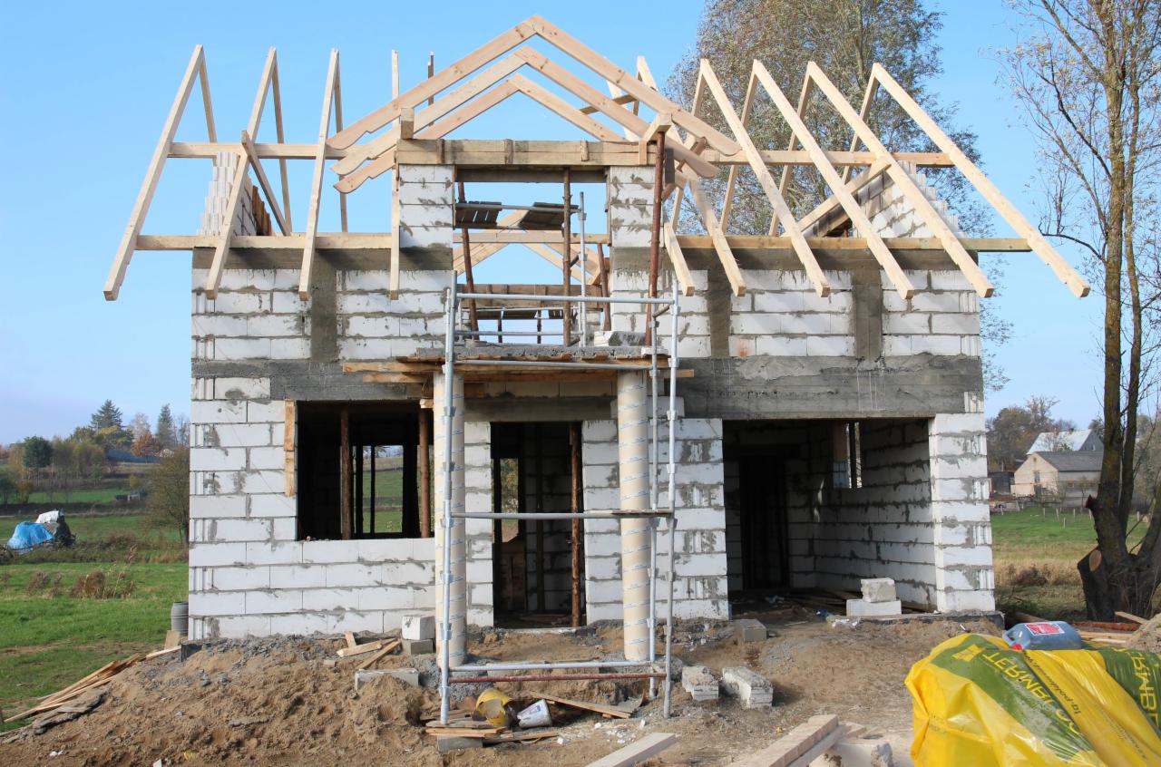 Surowy stan domu z bloczków betonowych w trakcie budowy, widoczne drewniane elementy konstrukcji dachu i rusztowanie, w tle krajobraz wiejski.