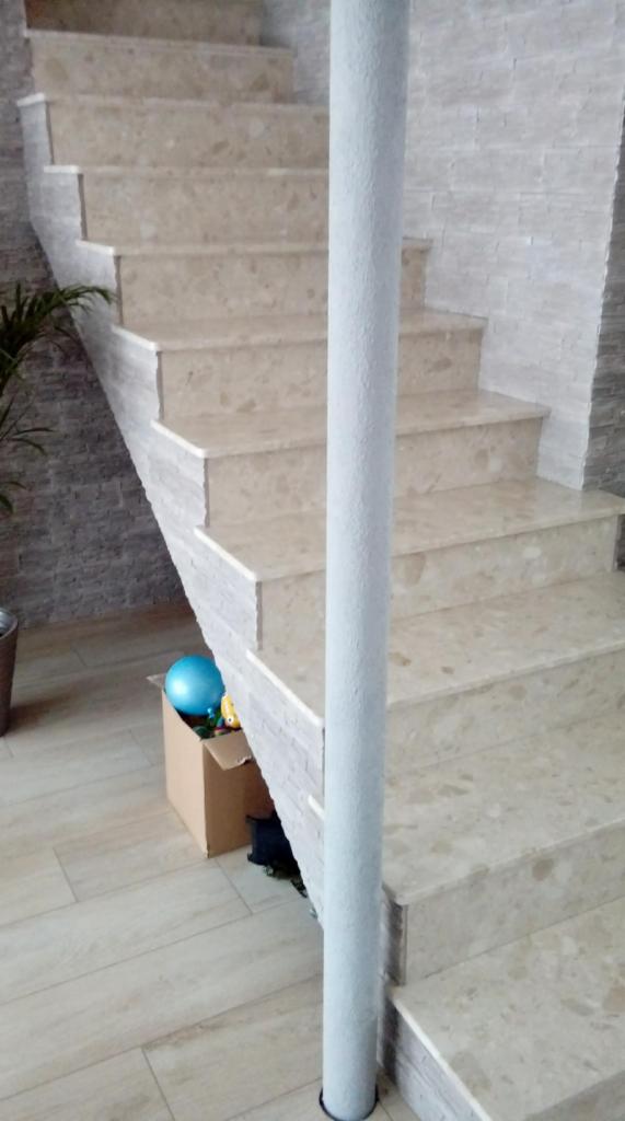 Marmurowe schody wewnętrzne z szarą kolumną wspierającą, widoczna ściana wykończona kamieniem dekoracyjnym i podłoga z paneli, w tle karton z zabawkami.