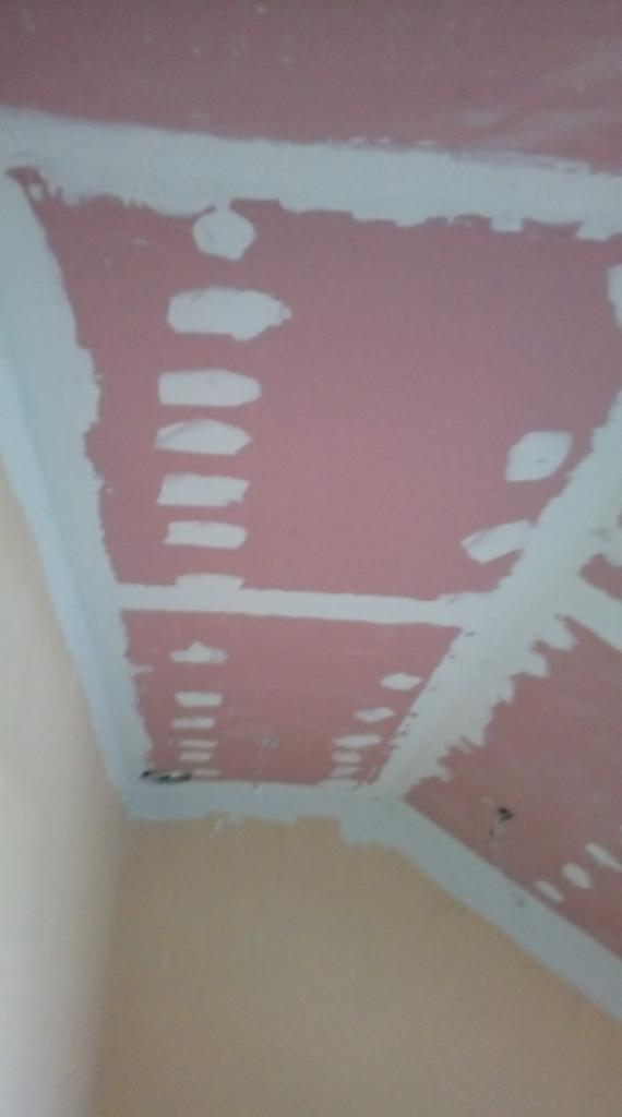 Różowa płyta gipsowo-kartonowa na suficie z widocznymi białymi paskami szpachli i otworami, przygotowana do dalszej obróbki na tle beżowej ściany.