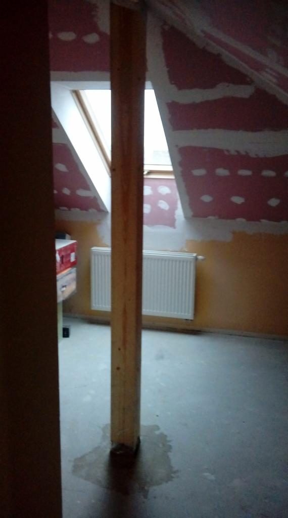 Poddasze w trakcie adaptacji: widoczny fragment ściany z płytami gipsowo-kartonowymi, grzejnik oraz drewniany słup podtrzymujący konstrukcję dachu, zacieki na podłodze.