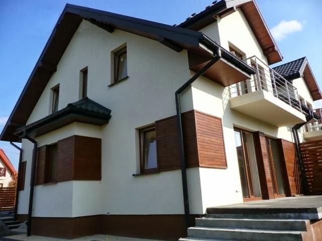 Dwukondygnacyjny dom z balkonem, jasną elewacją i elementami z drewna, widoczne rynny i schody wejściowe.