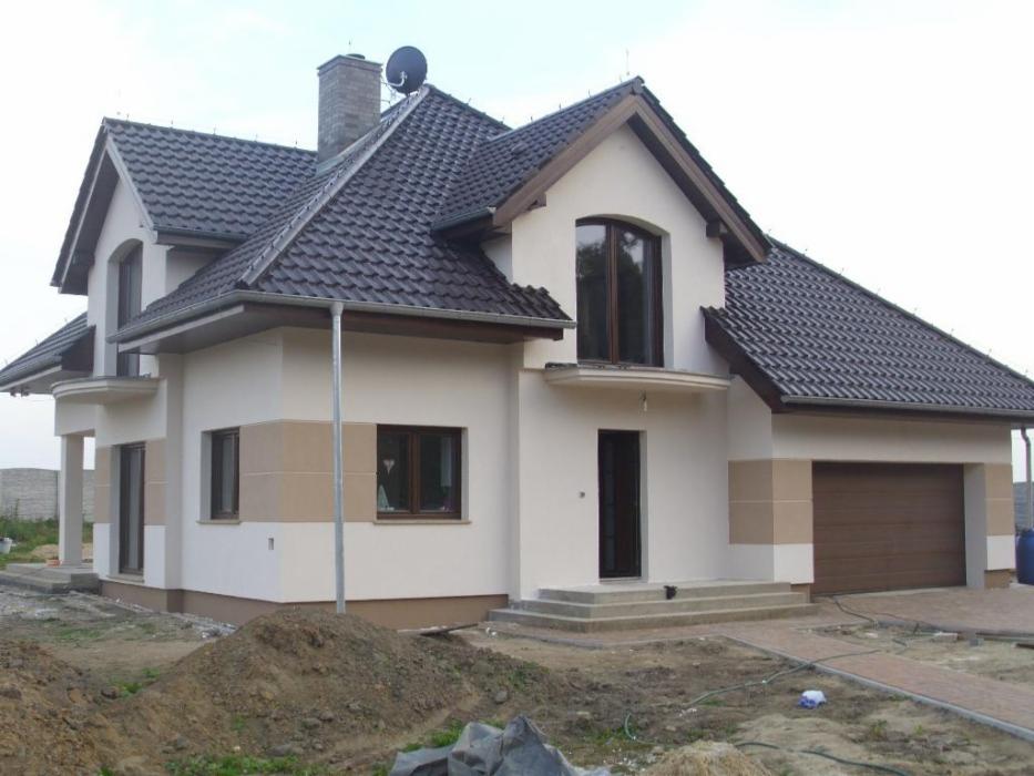 Nowoczesny dom jednorodzinny z ciemną dachówką, jasną elewacją z beżowymi akcentami, widocznym kominem i anteną satelitarną na dachu, z garażem po prawej stronie.