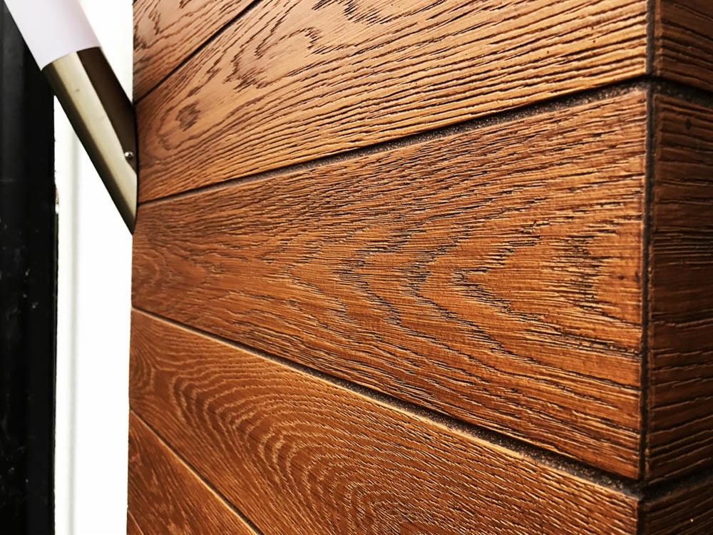 Fragment elewacji domu z brązowych, poziomych desek imitujących drewno, oświetlony lampą zewnętrzną, widoczny narożnik budynku.