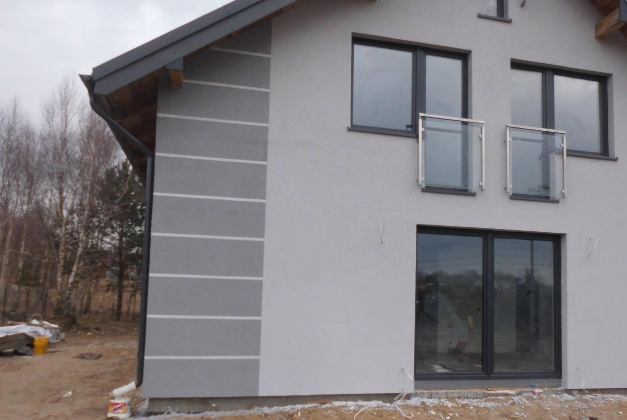 Dwukondygnacyjny dom murowany z szarą elewacją, poziomymi pasami dekoracyjnymi i małymi balkonami z szklanymi balustradami.